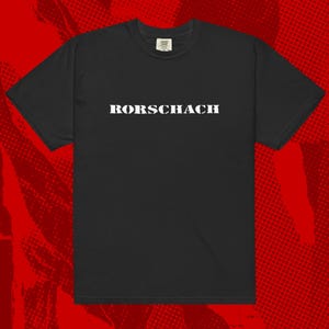 May include: Black t-shirt featuring the word "RORSCHACH" in bold white capital letters. The shirt has a classic crew neck and short sleeves. The background is a red halftone pattern.