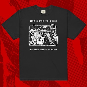 Puede incluir: Camiseta negra con texto blanco que dice "His Hero Is Gone" y un gráfico de una gasolinera con una persona de pie delante. El texto debajo del gráfico dice "Fifteen Counts of Arson."