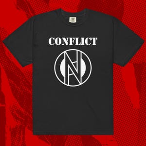 Może przedstawiać: Czarny t-shirt z napisem "CONFLICT" w białych literach nad białym okrągłym wzorem ze stylizowanym "N" w środku. Koszulka ma krótkie rękawy i okrągły dekolt.
