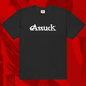 May include: Black t-shirt with the word "Assück" in white, stylized lettering across the chest. The shirt has a classic crew neck and short sleeves. The background is a red halftone pattern.