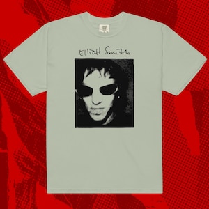 May include: A light green t-shirt featuring a black and white portrait of a person wearing sunglasses. The name "Elliott Smith" is written above the portrait in black cursive script. The shirt has a crew neck and short sleeves.