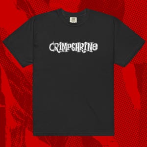 May include: Black t-shirt with the white text "CRIMPSHRINE" across the chest. The text has a decorative, vintage-style font. The shirt is a crew neck and appears to be made of a soft material.