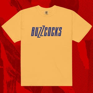 May include: Mustard yellow t-shirt with the word "BUZZCOCKS" printed in blue across the chest. The shirt has short sleeves and a crew neck. The background is a red halftone pattern.
