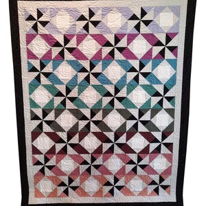 May include: A handmade quilt with a geometric pattern in shades of purple, teal, and coral, accented with black and white. The quilt features a black border and intricate quilting details, showcasing a blend of colors and textures.