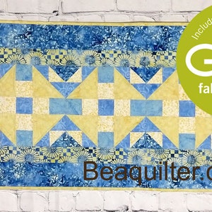 Peut inclure: Une courtepointe bleue et jaune avec un motif géométrique. La courtepointe est encadrée d'une bordure de fleurs jaunes et blanches. La courtepointe est étiquetée "Beaquilter.com" et porte un logo "GO!" avec le texte "fabric cutter friendly".
