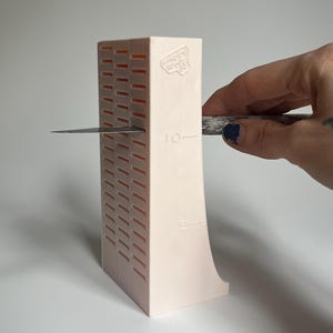 May include: A pink plastic knife sharpener with a grid pattern and markings for 5 and 10. A hand is holding a knife against the sharpener.