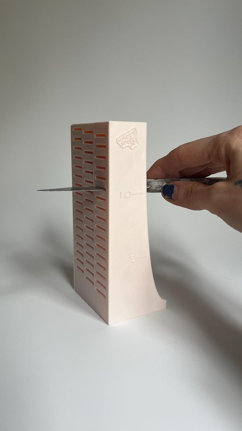 May include: A pink plastic knife sharpener with a grid pattern and markings for 5 and 10. A hand is holding a knife against the sharpener.
