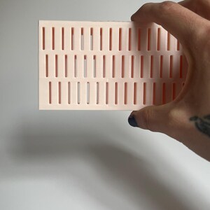 May include: A pink plastic grid with rectangular holes. The grid is being held by a hand.