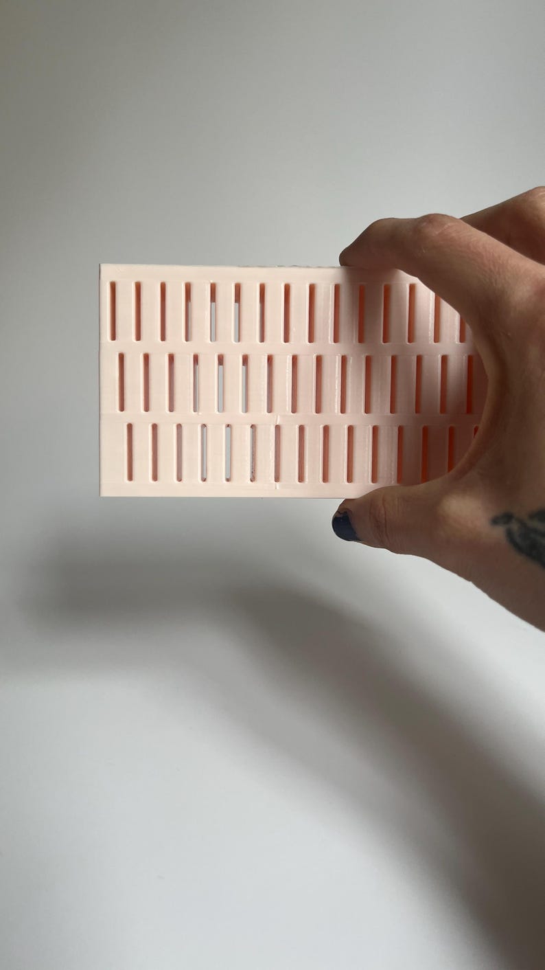 May include: A pink plastic grid with rectangular holes. The grid is being held by a hand.