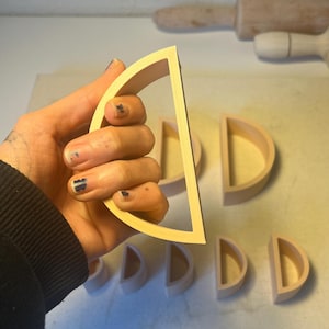 May include: A hand holds a light-colored wooden semi-circle shape, with several others arranged on a light-colored surface. A rolling pin is visible in the background. The shapes appear to be craft or decorative items.