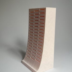 May include: A light pink 3D printed model of a building with a curved facade and a grid pattern.