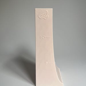 May include: A white plastic measuring tool with a curved edge and markings for 5 and 10. The tool has the words "Gooey Tools" embossed on the top.