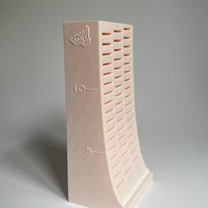 May include: A pink plastic tool for making clay beads. The tool has a curved shape and a series of slots for creating different sizes of beads. The tool is marked with the numbers "5" and "10" and the words "Good Tools".