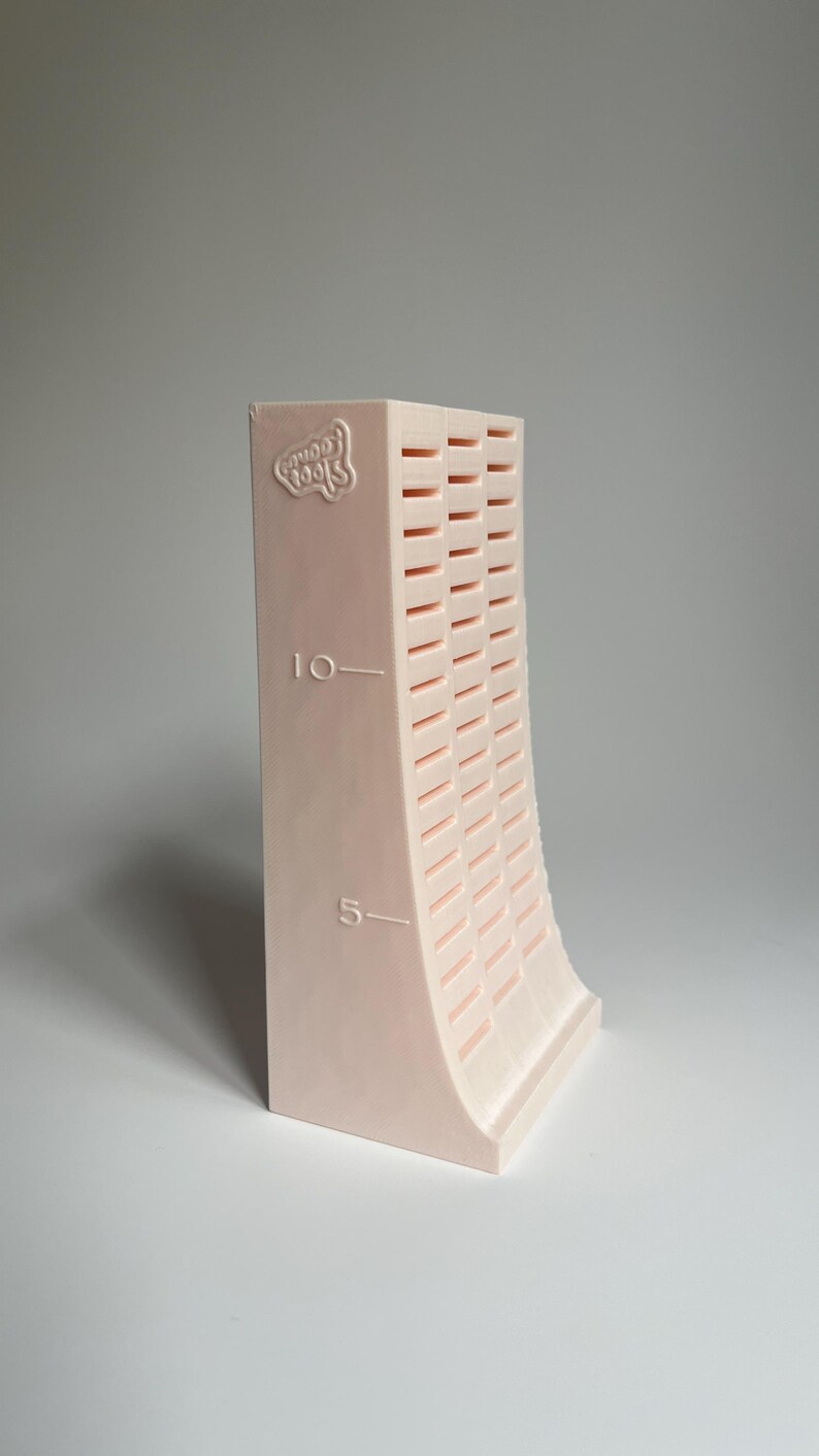 May include: A pink plastic tool for making clay beads. The tool has a curved shape and a series of slots for creating different sizes of beads. The tool is marked with the numbers "5" and "10" and the words "Good Tools".