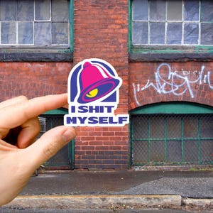 Puede incluir: Una pegatina con una parodia del logotipo de un restaurante de comida rápida. El logotipo incluye una campana rosa, un contorno azul y el texto "I SHIT MYSELF" en blanco. La pegatina se sostiene frente a una pared de ladrillos con ventanas.