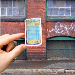 May include: A yellow and blue guest check with the number 59171 printed on it. The check is held in a hand against a brick wall background.