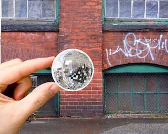 May include: A silver disco ball sticker with a white border. The sticker is being held by a hand in front of a brick wall with two windows and a green metal door.