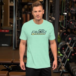 May include: A light green t-shirt with the text "Baja Blessed" in a stylized font. The shirt is short-sleeved and appears to be made of a soft material. The background is a gym setting with exercise equipment.