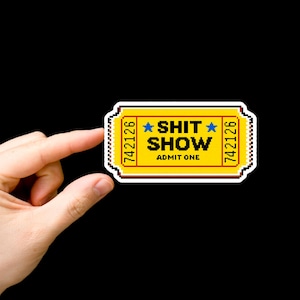 May include: A yellow ticket-shaped sticker with the words "SHIT SHOW" and "ADMIT ONE" in a pixelated font. The ticket has a white border and the number "742126" on each side. The background is black.