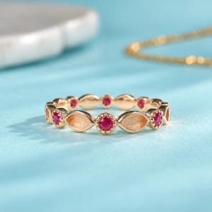 May include: A gold band ring featuring an alternating pattern of round, vibrant red gemstones and smooth, marquise-shaped gold settings. The ring has a polished finish and a continuous design that encircles the entire circumference of the band.