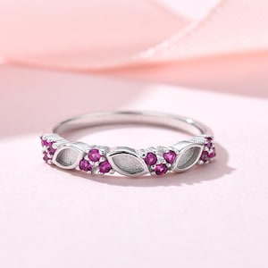Könnte beinhalten: Ein silberfarbener Ring mit einem sich wiederholenden Muster aus kleinen, runden, leuchtend pinkfarbenen Steinen in Dreiergruppen, die sich mit glatten, blattförmigen Metallaussparungen entlang der Ringschiene abwechseln.