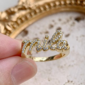 Puede incluir: Un anillo dorado con el nombre "mila" escrito en cursiva. Las letras están incrustadas con pequeñas piedras preciosas transparentes. El anillo se muestra sobre un fondo borroso, sugiriendo un tema de joyería o moda.