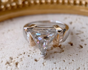 Pode incluir: Anel de prata com uma grande pedra preciosa transparente com corte triangular. O anel tem detalhes dourados e uma banda de prata. A pedra preciosa reflete várias cores e está sobre uma superfície texturizada.
