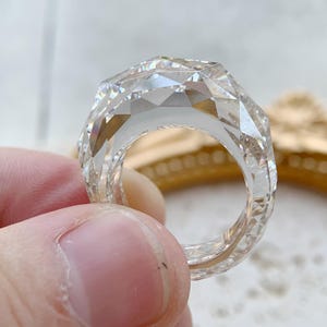 Könnte beinhalten: Ein klarer, facettierter Ring mit geometrischem Design. Der Ring wird von den Fingern einer Person gehalten. Die Facetten des Rings fangen das Licht ein und erzeugen einen funkelnden Effekt. Der Ring scheint aus einem transparenten Material zu bestehen.