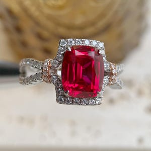 May include: A close-up of a ring featuring a large, rectangular, vibrant red gemstone. The gemstone is surrounded by a halo of small, clear stones. The band is silver with rose gold accents.