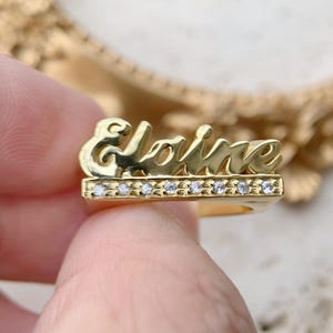 May include: A gold-toned ring featuring the name Elaine in a cursive script font. Below the name, a horizontal bar is set with eight small, clear, round-cut stones. The ring has a polished, reflective finish and a structured, layered design.