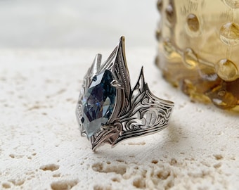 May include: A silver-toned ring with an elaborate, gothic-inspired design. The ring features a large, marquise-cut, blue-grey gemstone. Intricate patterns and pointed elements adorn the band, creating a regal and detailed aesthetic.