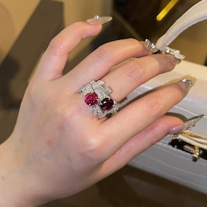 May include: A silver ring featuring two large, oval-cut, red gemstones. The ring is embellished with smaller, clear gemstones. The ring is on a hand with long, manicured nails.