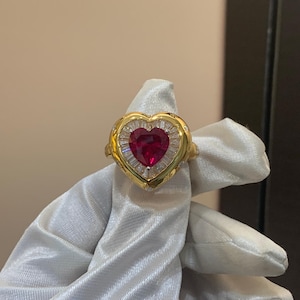 Könnte beinhalten: Ein goldener Ring mit einem herzförmigen Rubin-Mittelstein, umgeben von Diamanten im Baguetteschliff. Der Ring hat eine detaillierte Goldeinfassung und wird auf einer weißen Oberfläche präsentiert.