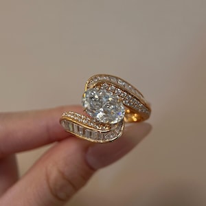 Puede incluir: Un anillo de oro con un gran diamante de talla ovalada como pieza central. El anillo está adornado con diamantes más pequeños y brillantes, creando un diseño lujoso y elegante. La banda se curva alrededor de la piedra principal, añadiendo un toque único.