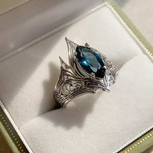 May include: A silver ring with an ornate design and a large, marquise-cut, teal-colored gemstone. The ring is set in a decorative setting with pointed edges, presented in a green and white ring box.
