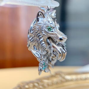 May include: A silver wolf head pendant with detailed fur and green gemstone eyes. The pendant is suspended, showcasing the side profile of the wolf's head. The pendant is intricately designed.