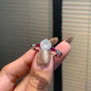 Op de afbeelding: Een zilveren ring met een grote, ronde, heldere edelsteen in het midden. De band heeft afwisselend rode en heldere edelstenen. De ring wordt vastgehouden door een hand met gouden glitter nagellak.