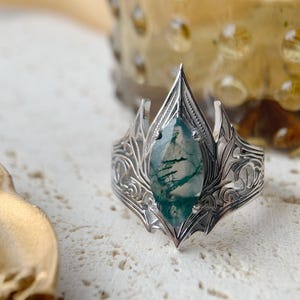 May include: A silver ring with an ornate, floral design. The ring features a large, oval-shaped, green and white moss agate stone. The ring's setting has a pointed top and detailed filigree work. The ring is on a textured, off-white surface.