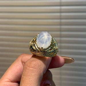 Op de afbeelding: Een gouden ring met een ovale, lichtgekleurde edelsteen met een stervormig patroon. De ring heeft ingewikkelde, sierlijke details rond de edelsteen en langs de band, met kleine accentstenen.