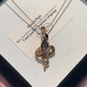 Puede incluir: Un collar con colgante de serpiente de color dorado con una piedra preciosa oscura y detalles en negro. La serpiente está enrollada alrededor de la piedra preciosa. El collar se exhibe en una caja negra con una etiqueta blanca con texto.