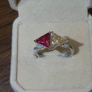 May include: A silver ring with two triangular gemstones, one red and one yellow, set side by side. The ring band has a twisted design and is encrusted with small, clear stones. The ring is displayed in a beige jewelry box.