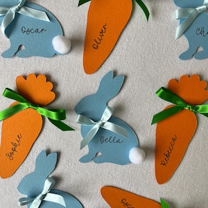 May include: A collection of Easter-themed place cards. Light blue bunny-shaped cards with white pom-pom tails and light blue ribbon bows. Orange carrot-shaped cards with green ribbon bows. Names are written on the cards.