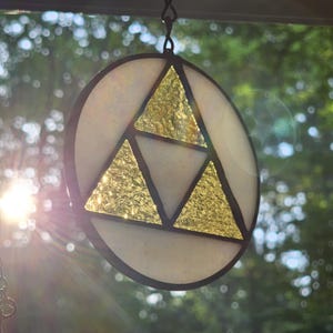 May include: A stained glass suncatcher featuring a circular design with a black border. The design includes three gold triangles and a white background. The suncatcher is hanging from a chain, with a blurred green background.