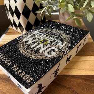 May include: A black book with a gold and white design on the cover. The book is covered in rhinestones and has the title "Court of Wings and Ruin" on the front. The book is sitting on a wooden surface.