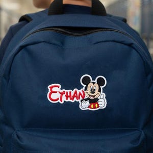Pode incluir: Uma mochila azul-marinho com um emblema bordado no bolso frontal. O emblema exibe o nome Ethan em letras cursivas vermelhas ao lado de um gráfico do Mickey Mouse fazendo um sinal de positivo, vestindo shorts vermelhos e luvas brancas.