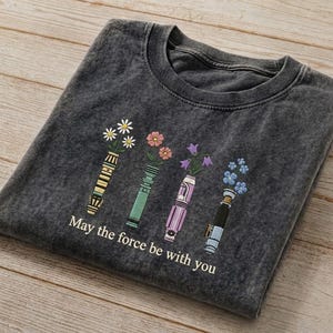 K&ouml;nnte beinhalten: Dunkelgraues T-Shirt mit floralem Design. Das Design zeigt vier stilisierte Lichtschwerter, die jeweils eine andere Art von Blume halten. Der Text "May the force be with you" ist darunter gedruckt.