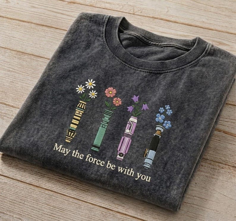 K&ouml;nnte beinhalten: Dunkelgraues T-Shirt mit floralem Design. Das Design zeigt vier stilisierte Lichtschwerter, die jeweils eine andere Art von Blume halten. Der Text "May the force be with you" ist darunter gedruckt.