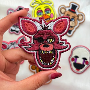 Puede incluir: Parches bordados con personajes de videojuegos. Los parches incluyen un zorro rojo con parche en el ojo, un pollito amarillo, un oso pardo con sombrero de copa y una calavera blanca con ojos morados. Los parches se sostienen en una mano.