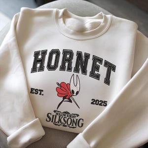 Puede incluir: Sudadera color crema con la palabra "HORNET" en letras negras. Debajo, un gráfico de un personaje con capa roja y aguja. Los textos "EST. 2025" y "SILKSONG" también están impresos en la sudadera.