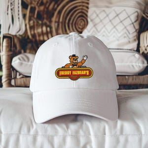 May include: White baseball cap featuring a cartoon bear with a top hat waving from a sign that reads "Freddy Fazbear's Pizza". The sign is outlined in gold with red and yellow accents. The cap is made of a soft, durable material.